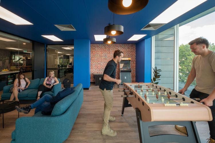 Employees relaxing and playing foosball in a bright office lounge with blue and brick walls.