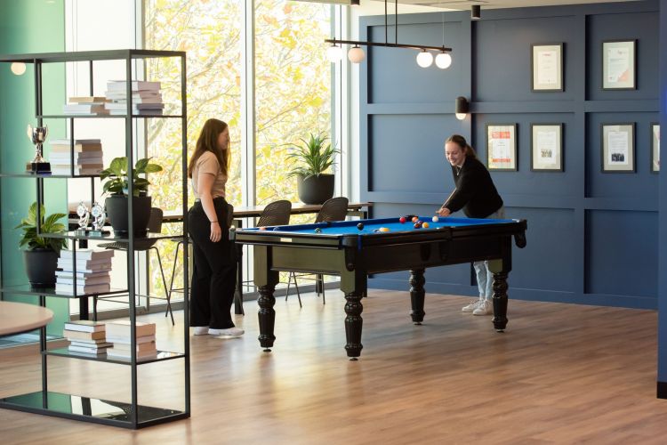 Two women playing pool in a well-lit office recreation area with blue walls and framed awards.