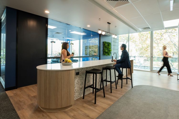 A modern office kitchenette with a round, wooden island countertop, black bar stools, and a wall-mounted green plant. 