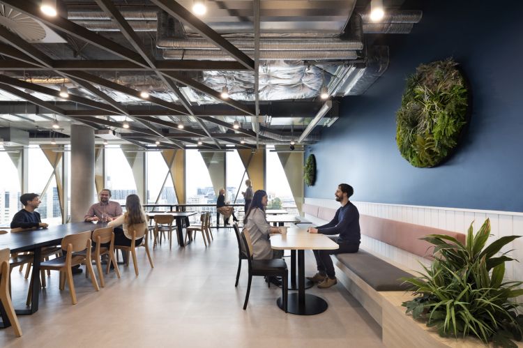 A spacious office break room with exposed ceiling ducts, wooden tables with matching chairs. Large windows offer a view of the cityscape. People are seen conversing and working.