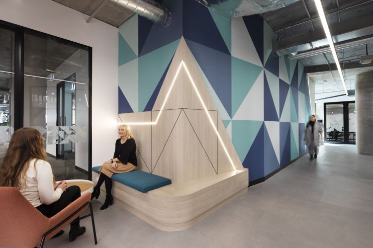 A modern office lounge area with geometric wall patterns in shades of blue, a bench with light wood paneling and blue cushion, and LED strip lighting. 