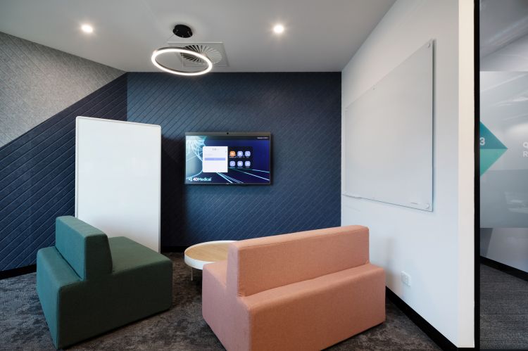 A modern office lounge with textured blue wallpaper, a green armchair, a coral-colored sofa, a round coffee table, and a digital screen on the wall.