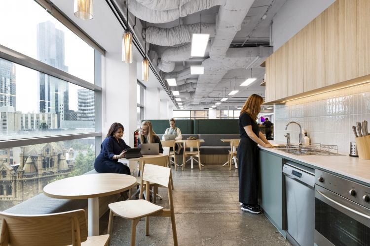 An office kitchen and dining area with a large window offering a city view. There's a kitchen counter with a sink, wooden tables and chairs, and people engaged in work and conversation. 