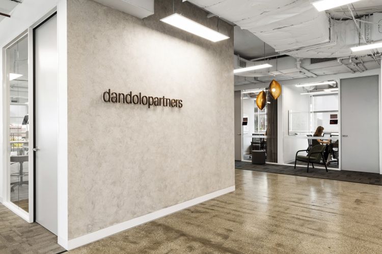 The entrance of an office with a textured wall featuring the 'dandolopartners' logo, a glass door to the left, and an open door to the right revealing a workspace with a person working at a desk