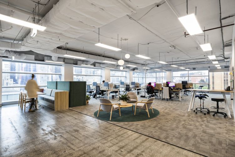 An expansive office floor with a mix of workstations, ergonomic chairs, a lounge area with a couch and armchairs, and a round table with chairs on a green rug.