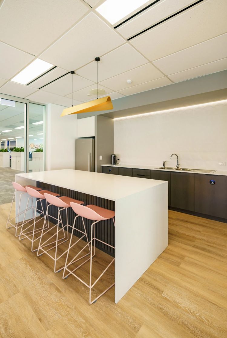 Modern office kitchen and breakout space with a white countertop island, pink stools, and a yellow pendant light.