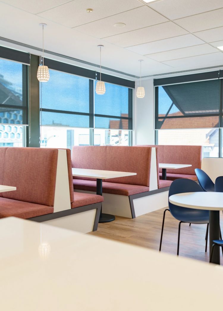 Office dining area with coral booths and white tables, blue chairs, and pendant lights, next to large windows with a city view