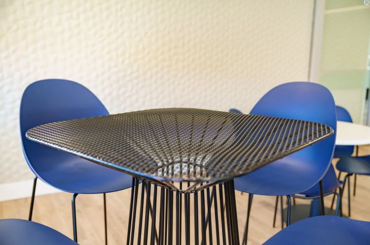 Close-up of a modern, wireframe table with a patterned surface, surrounded by blue shell chairs against a textured white wall.
