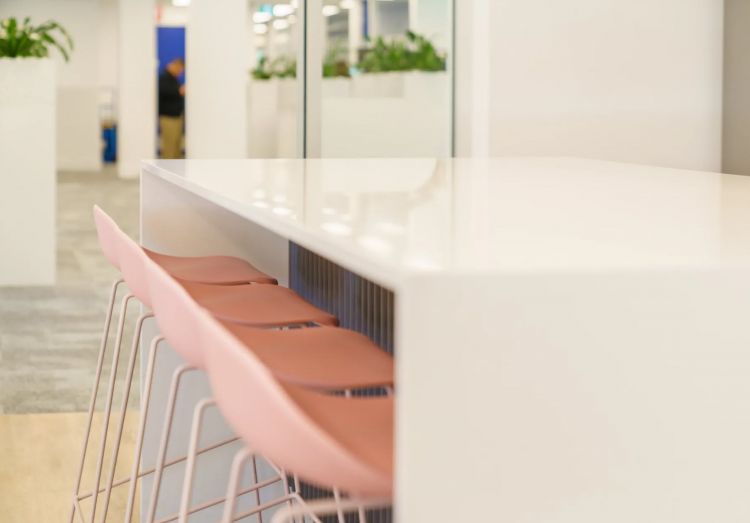 Close-up of a white countertop with pink chairs and metallic legs, with an office background