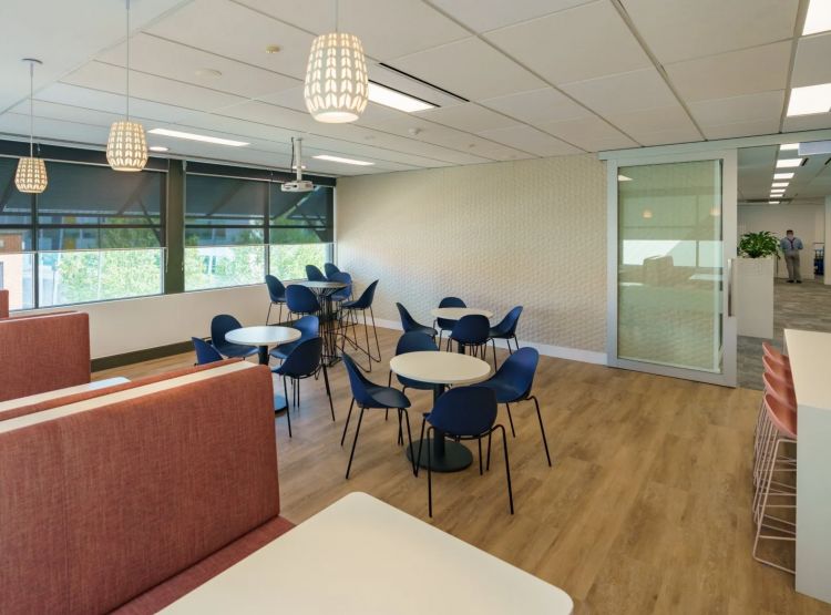 Open office area with blue chairs at white tables, pendant lighting, and glass-walled rooms in the background