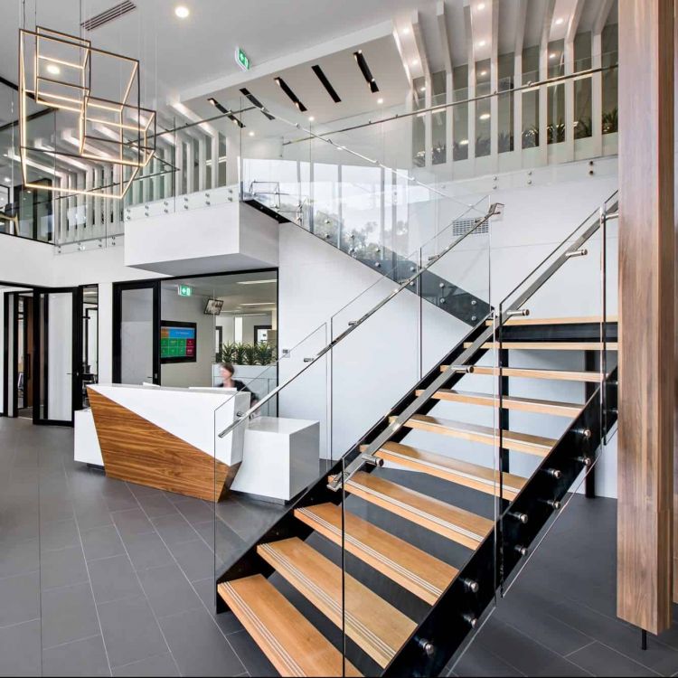 Modern office lobby with a sleek reception desk, wooden stairway with glass balustrades, and a minimalist chandelier.