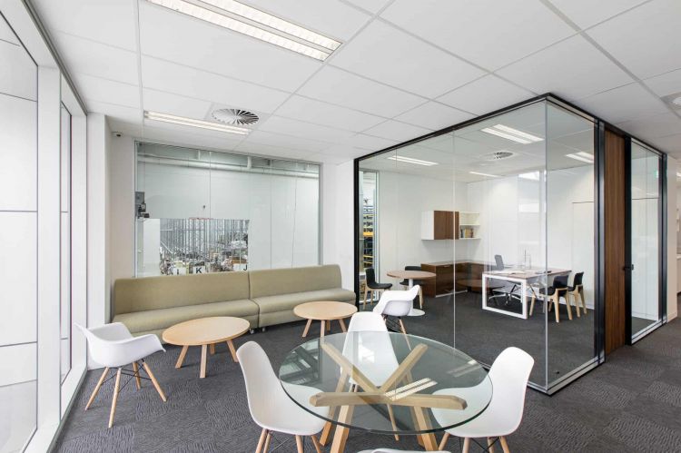 Bright office space with modern furniture, including a beige sofa, wooden tables, white chairs, and a glass-walled meeting room.