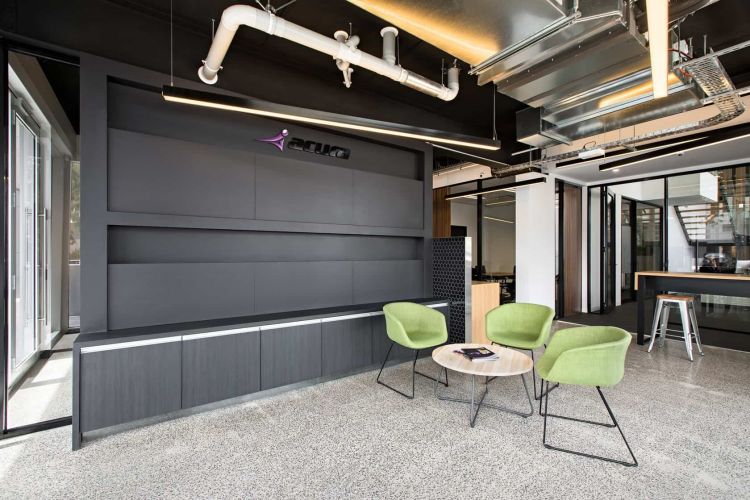 Industrial-style office breakout area with green chairs, a wooden table, and an Acura brand wall feature.