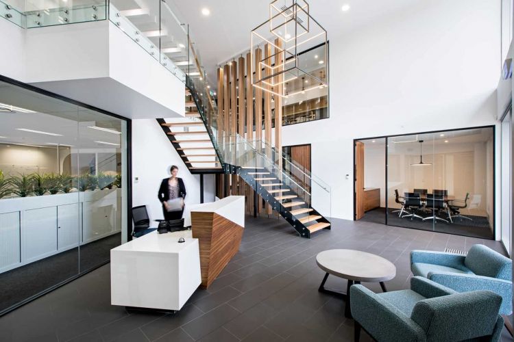 Spacious office lobby with modern furniture, a reception desk, a wooden and glass staircase, and seating areas.