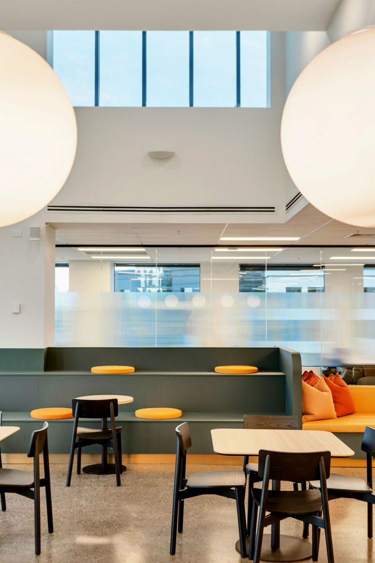 A bright office cafeteria with a minimalist style, large pendant lights, tall windows, and seating that includes dark chairs, light wood tables, and olive green booths with orange accents.