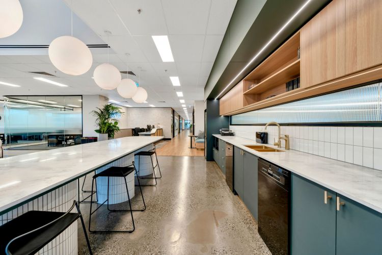 A spacious office kitchen with a long counter, black wire bar stools, globe pendant lights, wooden cabinetry, and a polished concrete floor.
