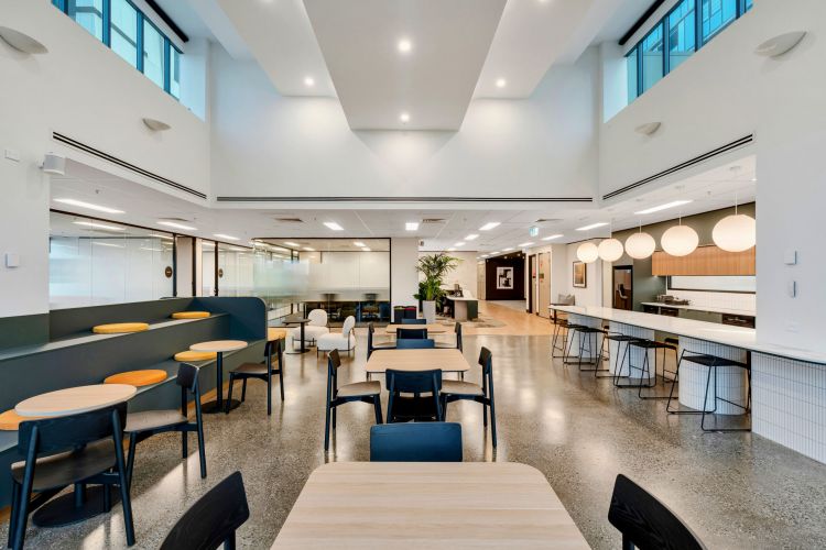 A modern, open-plan office dining area with natural light, featuring a mix of booth and table seating, large white globe lights, and an adjacent kitchen area with bar stools.