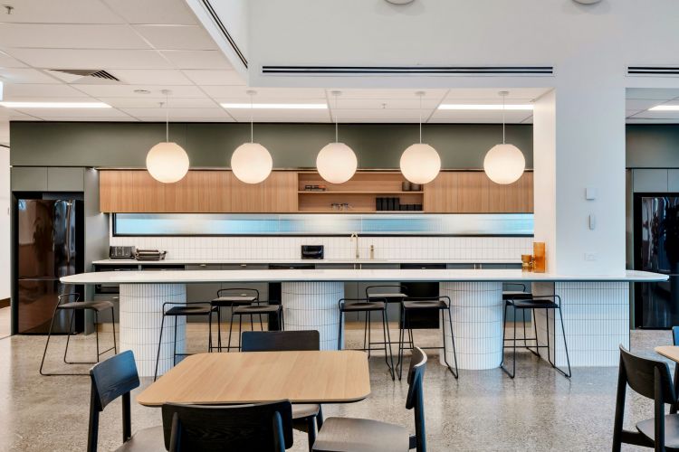 A modern office kitchen with a white tiled bar, black stools, wooden cabinetry, globe pendant lighting, and a communal dining area with tables and chairs.