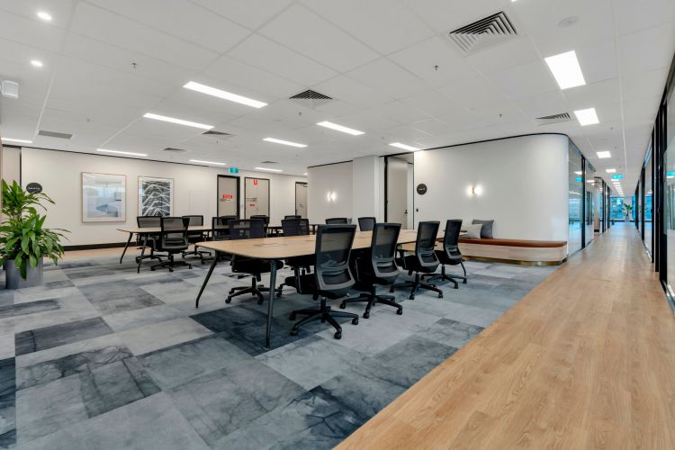 A modern office meeting room with a large table and chairs, patterned carpet, bright lighting, adjacent to a hallway with wood flooring and a casual seating nook with a bench and cushions.