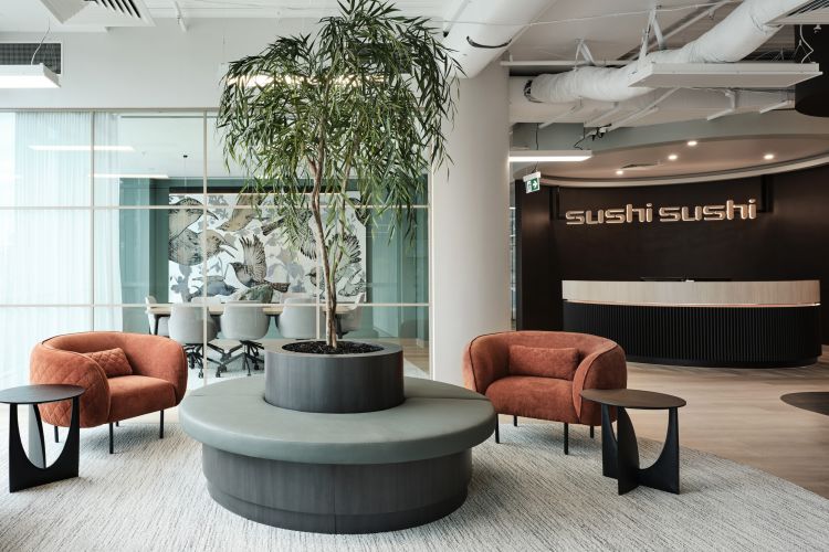 A modern office reception area with plush red chairs, a central tree planter, and a backdrop featuring the 'SUSHI SUSHI' logo.