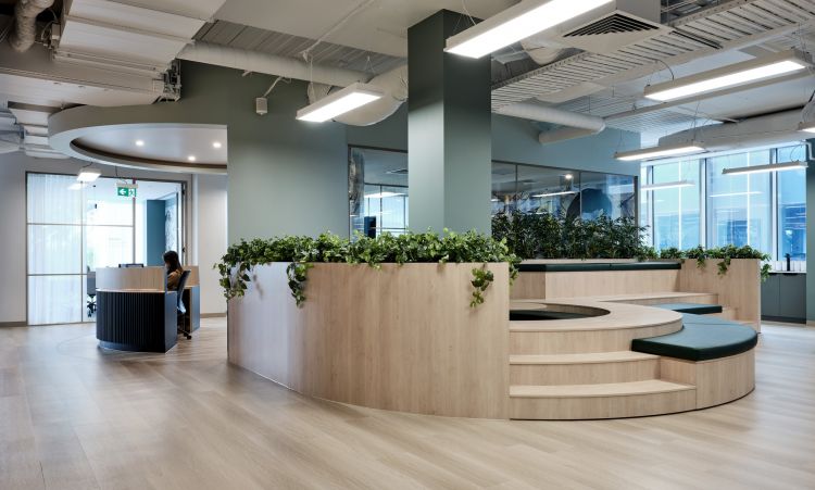 An office interior featuring circular wooden benches with planters, a reception desk, and large windows, in a spacious, well-lit room with a modern design.