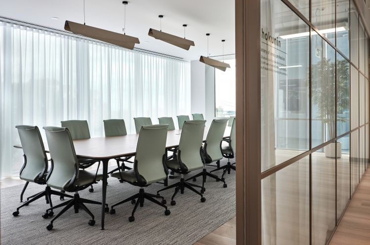 A modern conference room with a large table, green office chairs, and pendant lighting, viewed through glass walls.