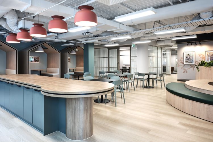  A spacious office cafeteria with round wooden tables, blue chairs, red hanging lamps, and booth seating under house-shaped structures.