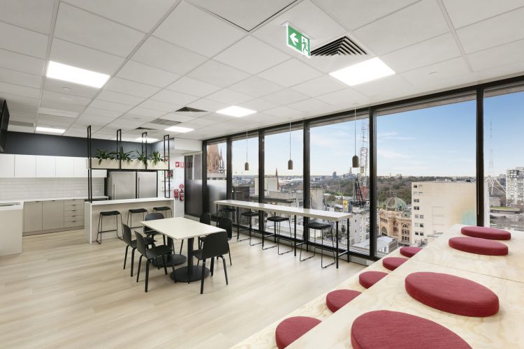 A bright office kitchen and dining area with large windows offering a city view, featuring modern furniture and pendant lights.
