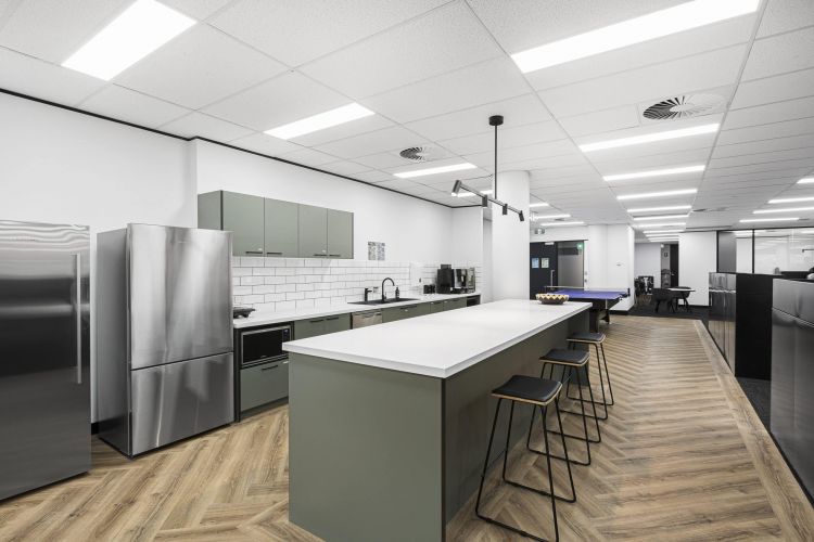 A modern office kitchen and break area with stainless steel appliances, white subway tile backsplash, gray cabinetry, and a long island with black stools on a herringbone pattern wooden floor. 