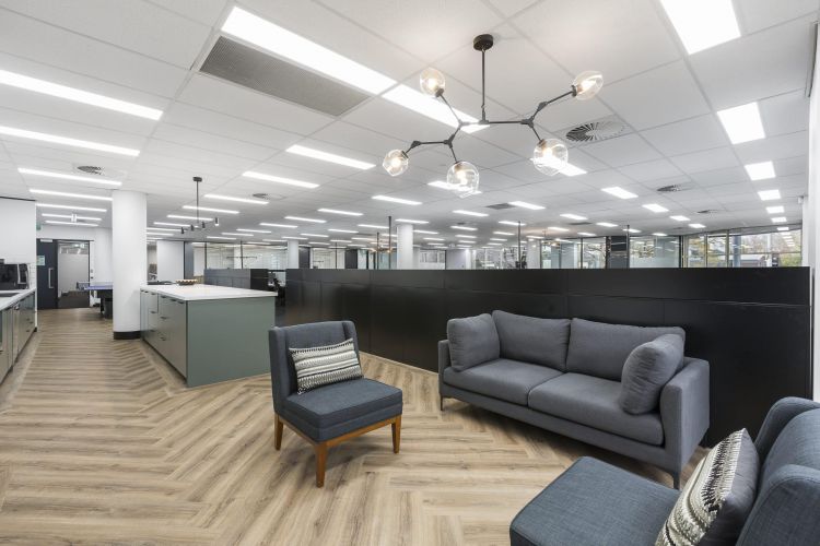 A spacious office lounge with contemporary gray sofas and armchairs, patterned cushions, a herringbone wooden floor, and a geometric light fixture.