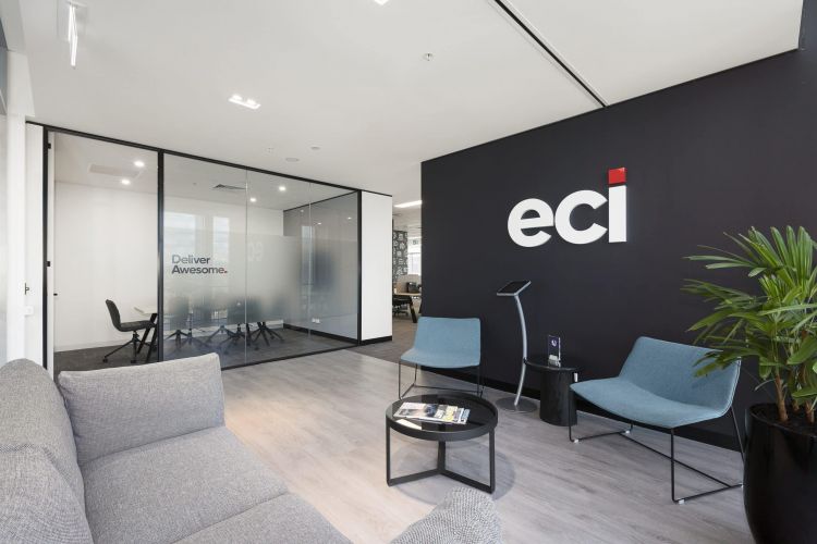 A cozy office lounge with gray sofas, blue chairs, a large 'eci' logo on the wall, and glass-walled meeting rooms in the background.